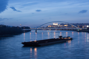 Old town, riverside, Bratislava castle, Apollo Bridge, UFO Bridge, New Bridge with cargo ship in foreground shoot during dusk from above river Danube, Slovakia
