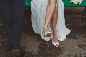 slender legs of the bride in white shoes with open nose and groom in a park on a bench in the summer