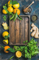 Ingredients for making natural hot drink with wooden tray in center. Oranges, mint, lemons, ginger, honey, apple over plywood background, top view, copy space. Clean eating, detox, dieting concept