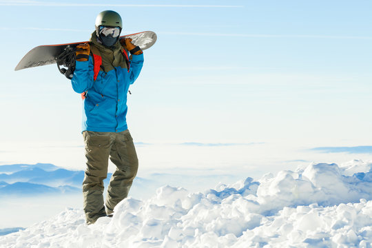 Young snowboarder in helmet at the very top of a snowy mountain with beautiful sky on background