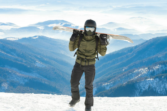 Young snowboarder holding snowboard on his sholders and walking to the very top of a mountain