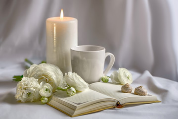 White objects on a white background. Burning candle, cup, notebook, cookies, ranunkulyus flowers.