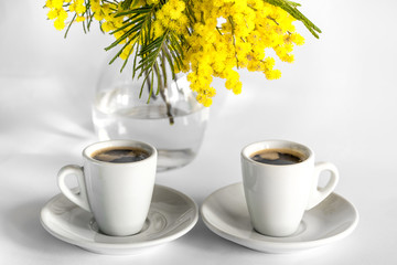 Two cups of coffee with plates and a vase from glass with branches of mimosa on a white background