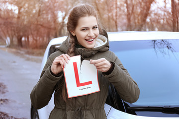 Happy young woman tearing learner driver sign while standing near car outdoors