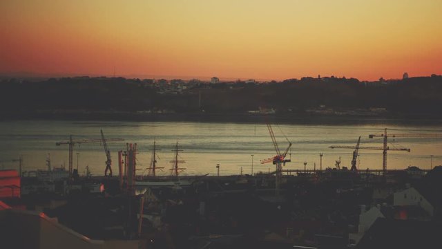 Time lapse from high above of nightfall on harbor, docks and river of evening European city with many working cranes, ships awash, floating barge, clear sky and residential district in foreground