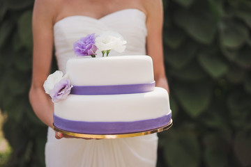 Beautiful slim caucasian woman holds a white wedding cake with purple flowers decoration outdoors on the background of green leaves