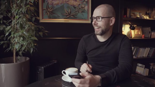 A young man in a cafe sitting at the table. He read a message on his mobile phone and smiling.