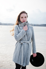 Young model girl in gray coat and black hat  posed against freeze lake.