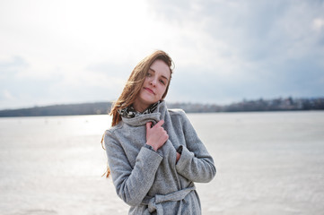 Young model girl in gray coat with red hair against freeze lake.