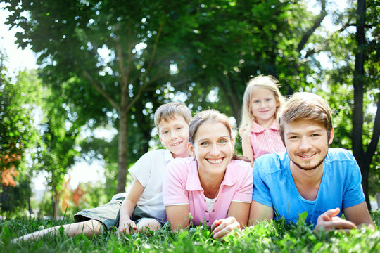young happy family at noon in the park on the grass. Two young parents and children, boy and girl, lies on the grass and smiling looking at the camera.