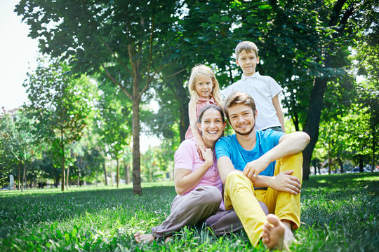 young happy family at noon in the park on the grass. Two young parents and children, boy and girl, sits on the grass and smiling looking at the camera.