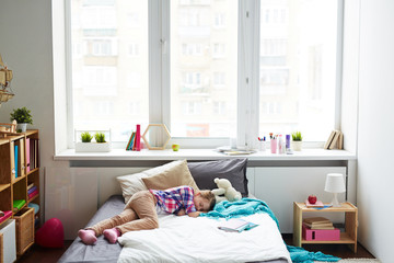 Little girl having a nap on her soft bed in bright modern room with large window