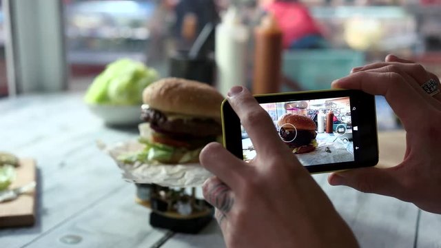 Cell phone photographing burger. Hamburger on table. Try food and share impressions.