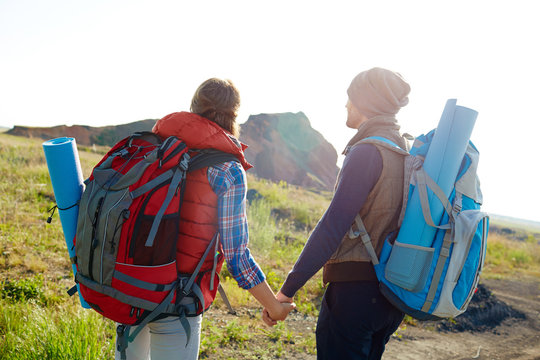 Rear view image of young tourist couple with big backpacks looking away holding hands walking to mountains together