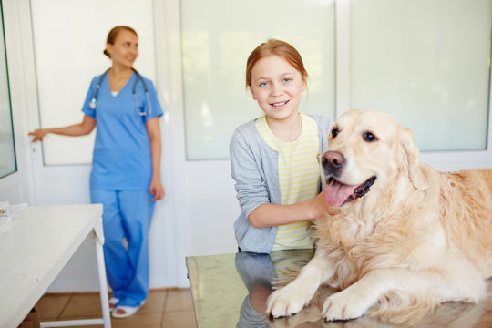 Small red-haired owner standing next to her dog resting on examination table after checkup and smiling at camera brightly