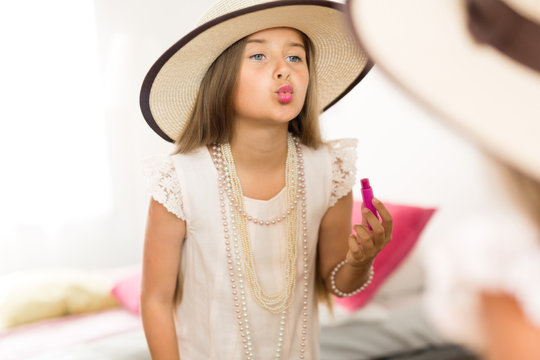 Mirror reflection shot of little girl playing dress up in mothers jewels and hat putting makeup and pink lipstick on, blowing air kisses and grimacing