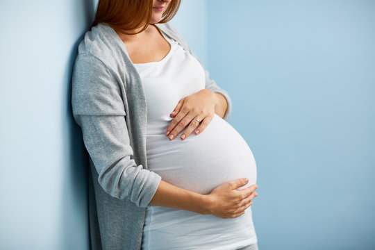 Mid-section of unrecognizable pregnant woman smiling and holding her big belly caringly against blue wall of childrens room