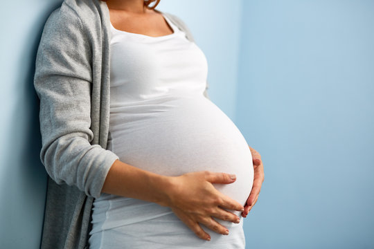 Mid-section portrait of unrecognizable woman during last months of pregnancy holding her big belly gently standing against wall in blue room