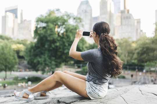 USA, Manhattan, young woman sitting in Central Park taking picture of skyline with smartphone