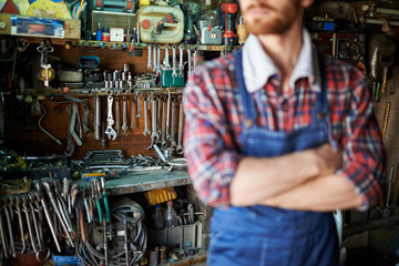 Closeup shot of blurred unrecognizable bearded male standing confidently with arms crossed against shelves of mechanics equipment and tools in garage workshop