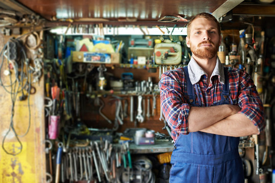 Portrait of handsome bearded mechanic standing confident with arms crossed  and looking at camera against shelves of tools and equipment in workshop