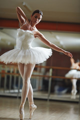Portrait of graceful ballerina wearing white tutu dancing in half-lit ballet studio practicing for...