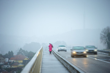 Storm weather. A woman in a pink coat with an umbrella is a sidewalk in the rain. Cars at high speed pass by.