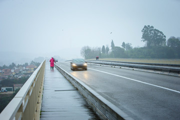 Storm weather. A woman in a pink coat with an umbrella is a sidewalk in the rain. Cars at high speed pass by.