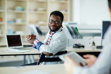Waist-up portrait of smiling bearded man sitting at desk in open plan office and asking his...