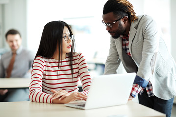 Fototapeta premium Beautiful Asian employee sitting at office desk and listening to her middle-aged senior with attention while he explaining her operating principle of computer program