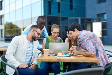 Multi-ethnic group of young creative people working together at outdoor table by modern building, looking at laptop screen and discussing ideas collaborating on project