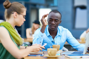 Portrait of two creative young business people working at outdoor area of modern office building: African-American man talking to busy girl beside him during coffee break