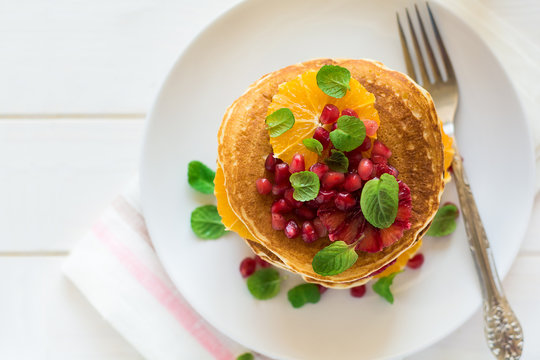 Traditional breakfast: stack of pancakes with orange slices and pomegranate seeds decorated mint leaves on white wooden table. Selective focus