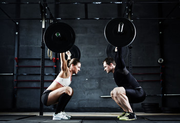Motivational wide shot of young man and woman holding huge heavy barbells overhead looking at each...