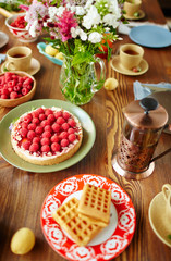 High-angle view of freshly baked raspberry tart, homemade waffles, summer flowers, French press and tea cups placed on wooden table