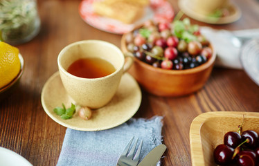 Cup of scented herbal tea on plate and bowls of ripe gooseberries and cherries on wooden background, close-up view