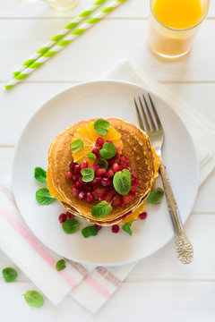 Traditional breakfast: stack of pancakes with orange slices and pomegranate seeds decorated mint leaves on white wooden table. Selective focus