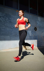 Young female athlete in short top running along the street, listening to music and smiling happily