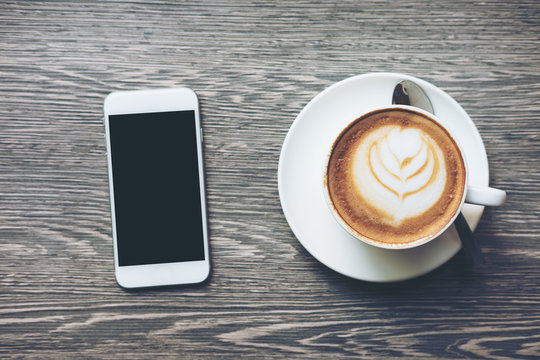 Mockup image of white mobile phone with blank black screen and hot latte coffee on vintage wooden table in cafe