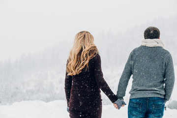 Look from behind at man and woman holding their hands together while they stand under falling snow