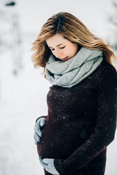 Thoughtful blonde woman looks at her pregnant belly posing outside under falling snow