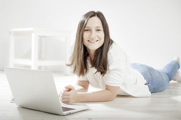beautiful young girl lying on the floor with a laptop and smiling, dressed in a white shirt with short sleeves and jeans