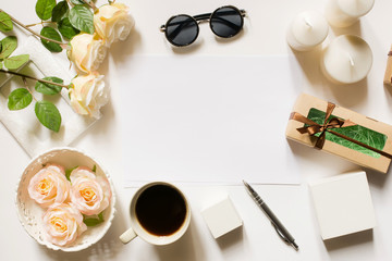 Obraz na płótnie Canvas Empty sheet with copy space on the white desk with coffee cup, sunglasses, roses, pen, gifts, candles and vintage white tray. Top view, flat lay, copyspace.
