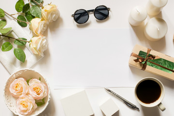 Obraz na płótnie Canvas Empty sheet with copy space on the white desk with coffee cup, sunglasses, roses, pen, gifts, candles and vintage white tray. Top view, flat lay, copyspace.