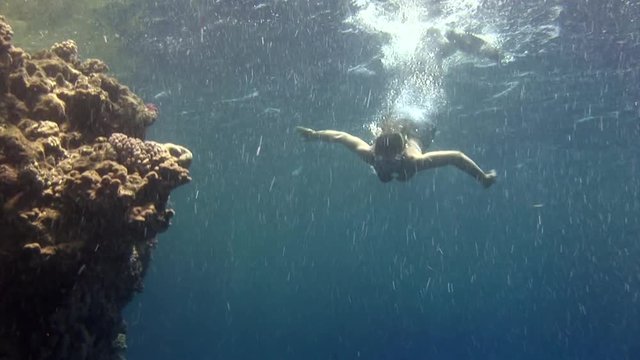 Underwater model free diver swims in clean transparent blue water in Red Sea. Young girl smiling at camera. Filming a movie in marine landscape, coral reefs, ocean inhabitants.
