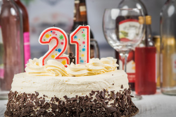 White cake with chooclate decorations and 21 candles.   Alcoholic beverages  blurred in background.