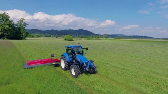 We can see a blue tractor driving on the field and it is cutting grass with red agricultural machinery. It is a nice summer day.
