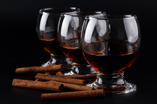 Three glasses with cognac in a row on a black background with cinnamon sticks. Selective focus and shallow depth of field