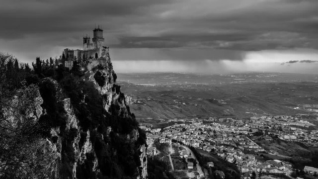 San Marino time-lapse, black and white. Fortress of Guaita on Mount Titano at sunset in San Marino. Time-lapse of heavy raining clouds with aerial view at the city. Car traffic lights and illumination