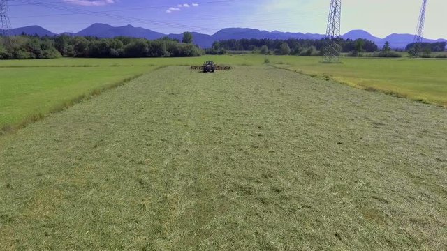 A tractor has to prepare a lot of hay on a big field. It is a beautiful summer day. Heli shot.
